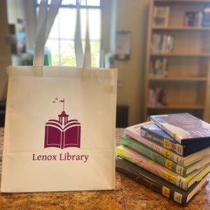 White tote bag with maroon Lenox Library logo next to a stack of books.
